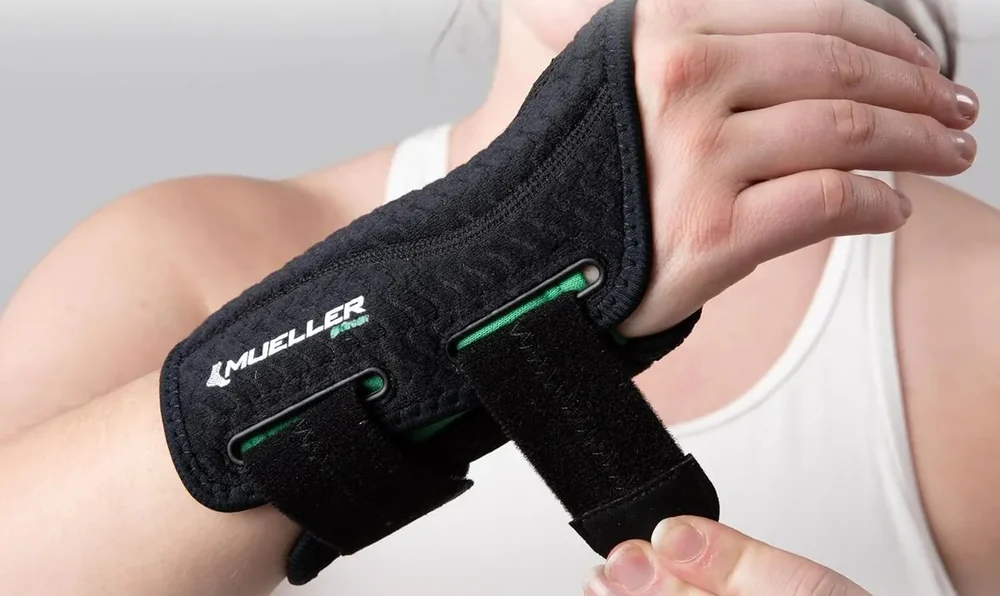 The Mueller Green Fitted Wrist Brace being tightened on a wearer's wrist, showing its two adjustable straps and green lining